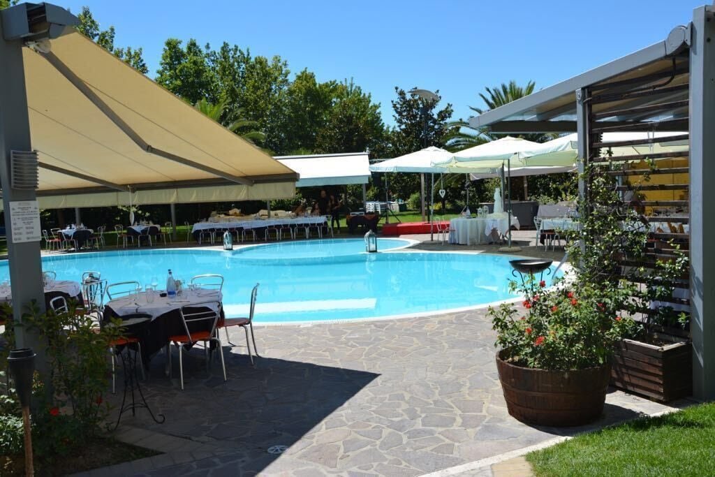 Una piscina con mesas y sillas alrededor, protegida por grandes sombrillas, y una planta en maceta cerca.