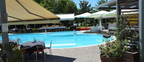 Una piscina con mesas y sillas alrededor, protegida por grandes sombrillas, y una planta en maceta cerca.