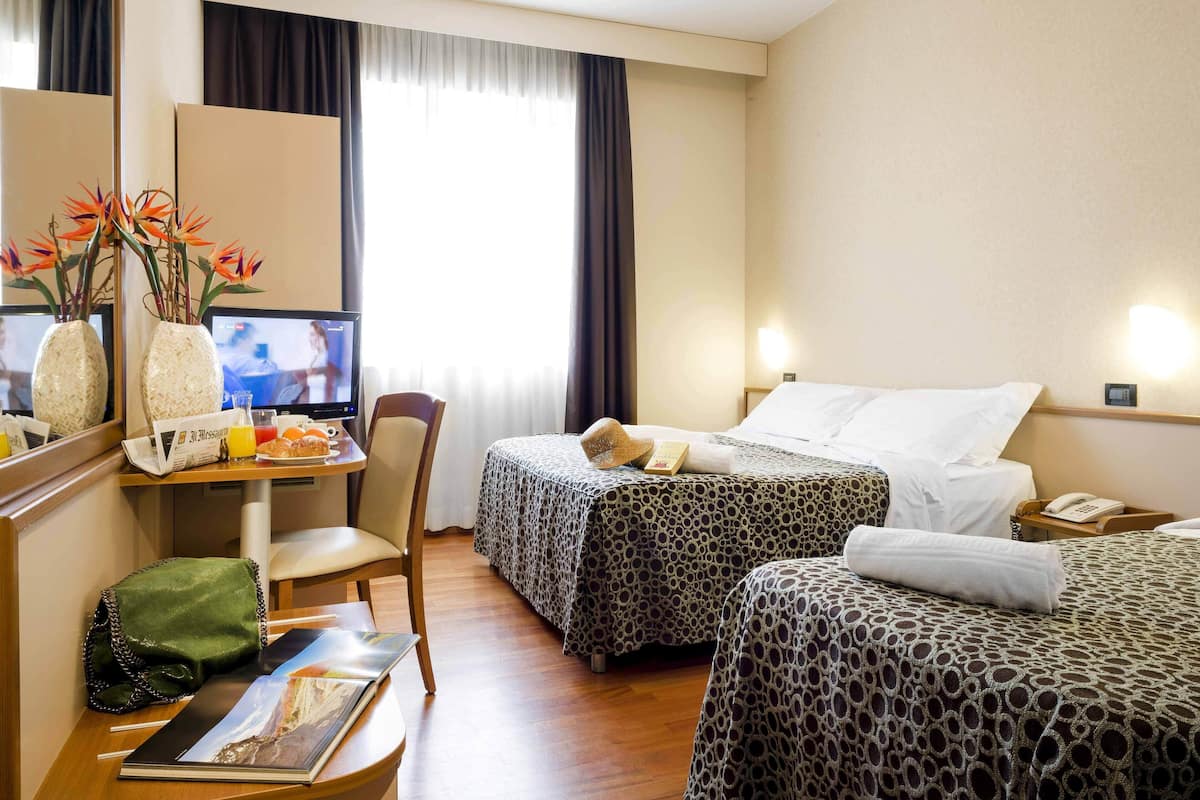 A hotel room with two beds, a desk with a computer, a chair, a green bag, and a vase with flowers.