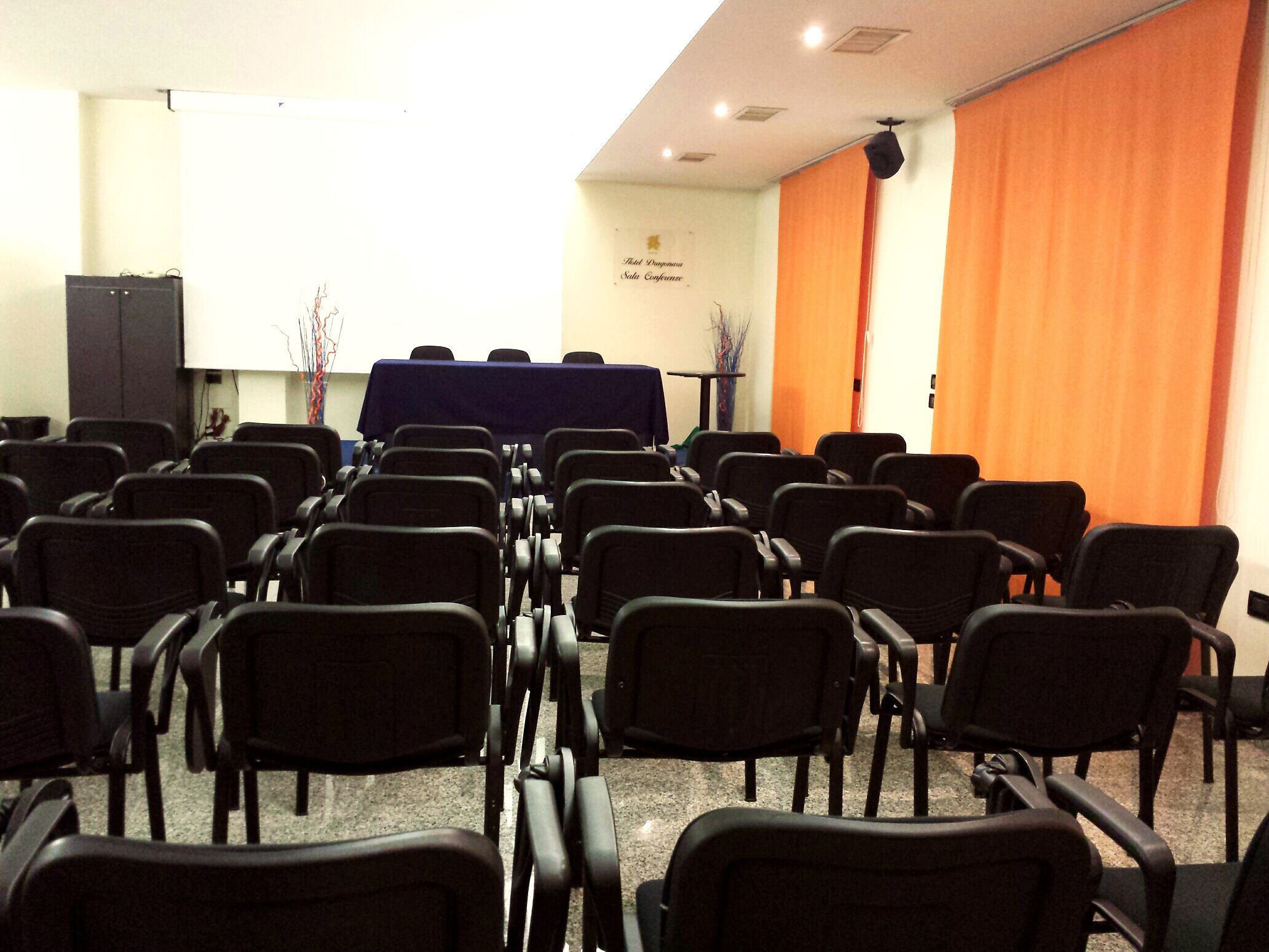 Ein Konferenzraum mit Reihen schwarzer Stühle, einem Tisch mit blauer Tischdecke und einem orangefarbenen Vorhang.