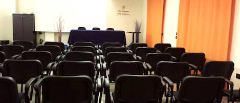 Un salón de conferencias con filas de sillas negras, una mesa con cubierta azul y una cortina naranja.