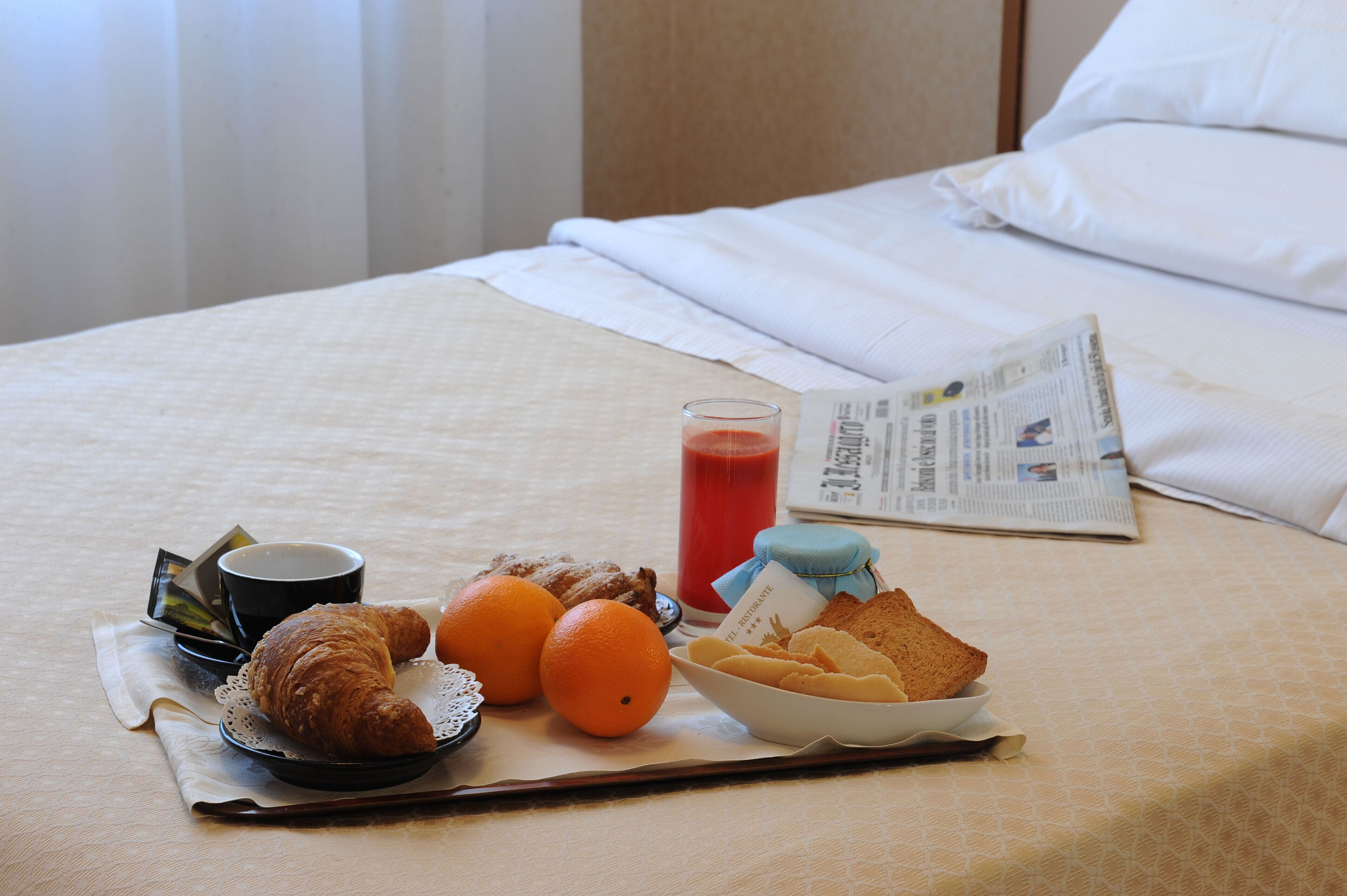 Ein ordentlich bezogenes Bett mit einer Zeitung, einem Glas rotem Saft, einem Croissant, zwei Orangen und einer Schale Kekse auf einem Tablett.