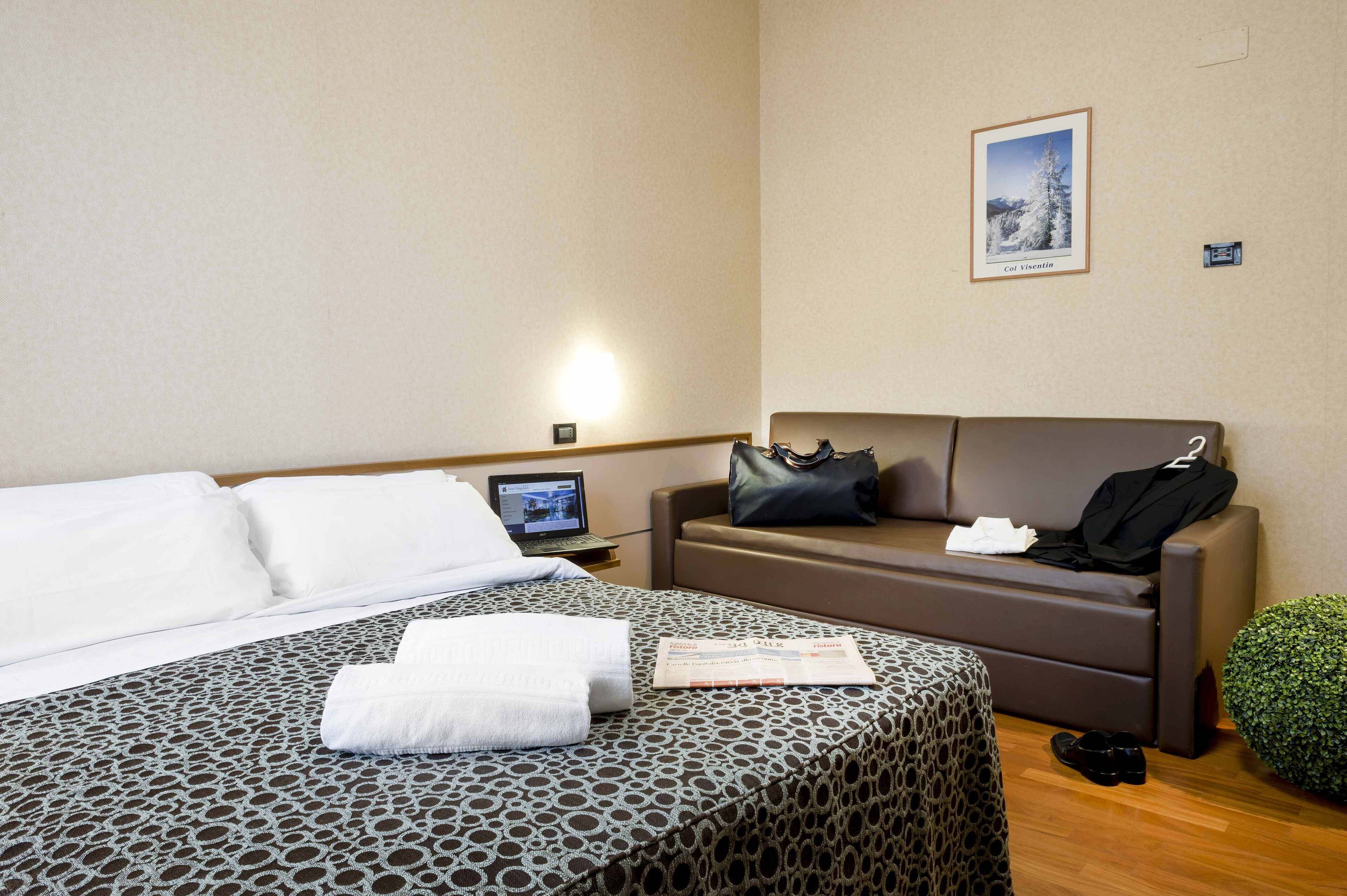 A hotel room with a bed, a sofa, a small table with a laptop, a framed picture on the wall, and a green plant.