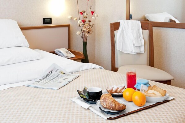 A neatly made bed with white linens, a bedside table with a lamp, a vase with dried flowers, and a breakfast tray with croissants, oranges, and a glass of red juice. - Dragonara (San Giovanni Teatino)