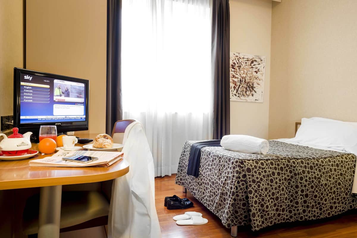 A hotel room with a bed, a desk with a computer, a window with curtains, and a wall art piece.