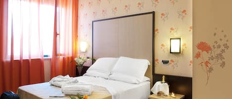 A hotel room with a bed, a nightstand, a lamp, a suitcase, and a window with curtains.