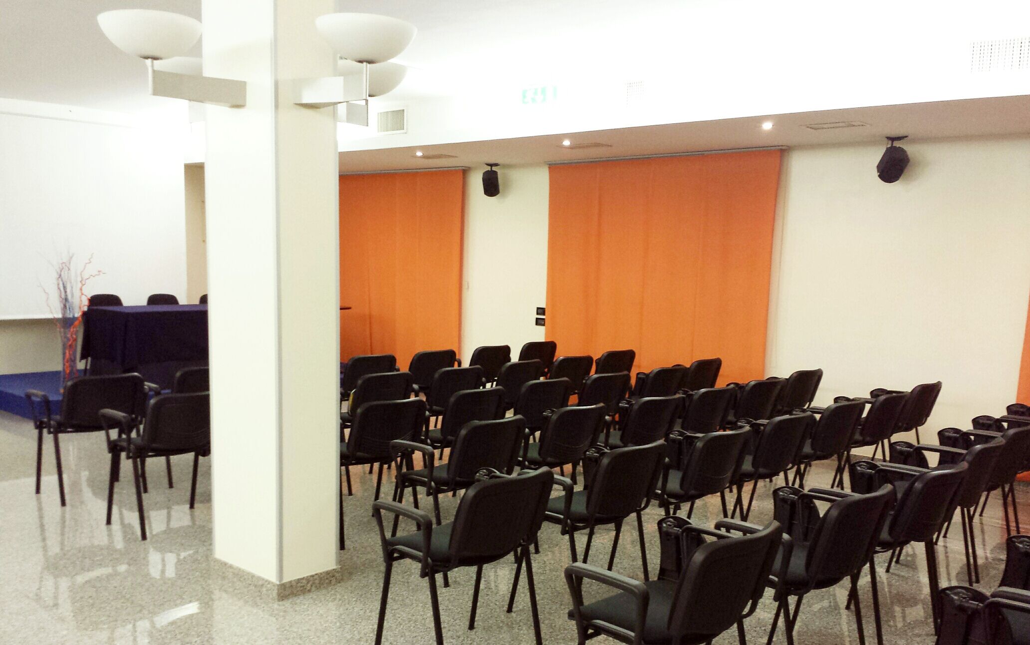 Un salón de conferencias con filas de sillas negras, un atril y cortinas naranjas.