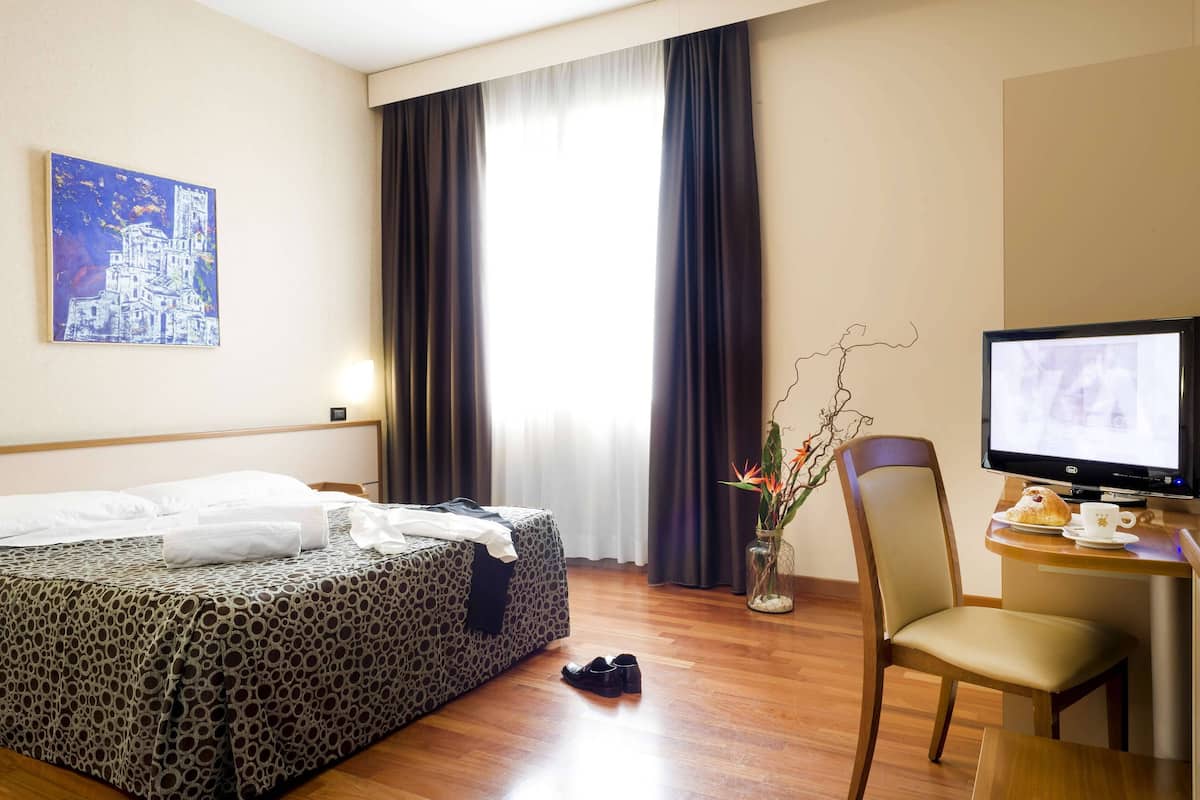 A hotel room with a bed, a desk with a computer, a chair, a vase with flowers, and a painting on the wall.