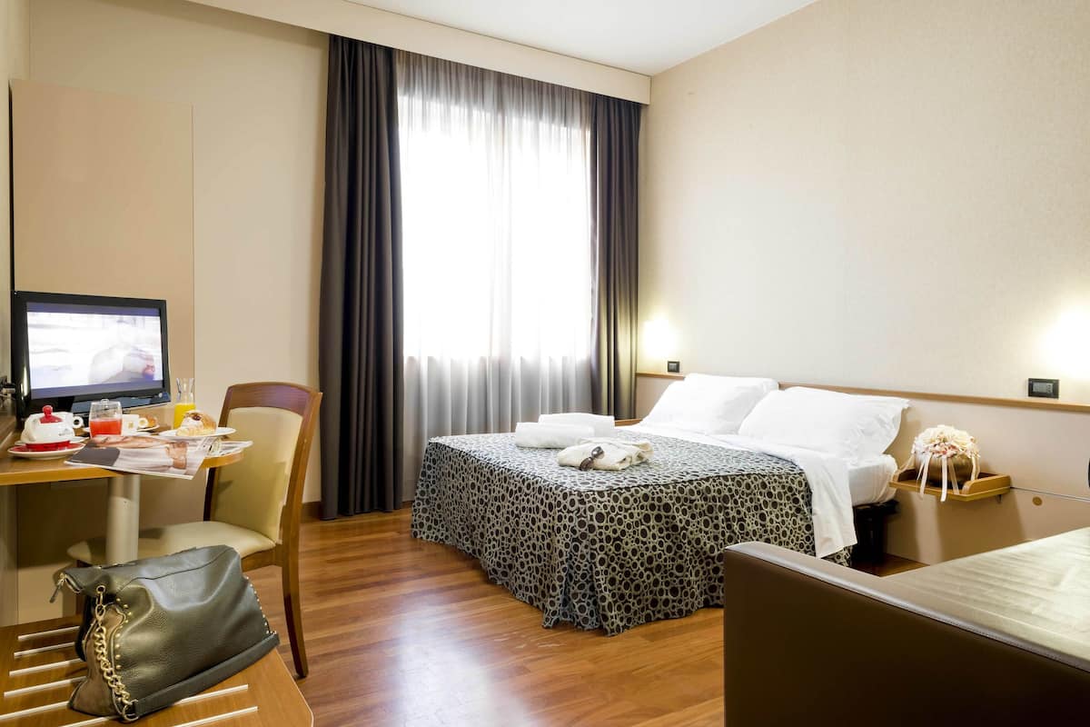A hotel room with a bed, a desk with a computer, a chair, a handbag, and a window with curtains.