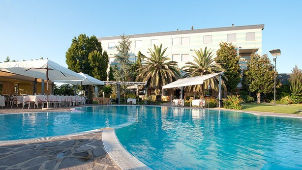 Una piscina con sillas de descanso y sombrillas, rodeada de palmeras y un edificio al fondo.