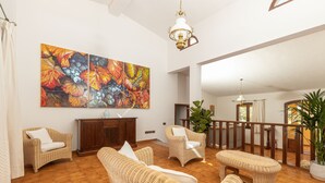 Uma sala ampla com móveis de vime, uma grande pintura na parede e um corrimão de madeira com vista para uma varanda.