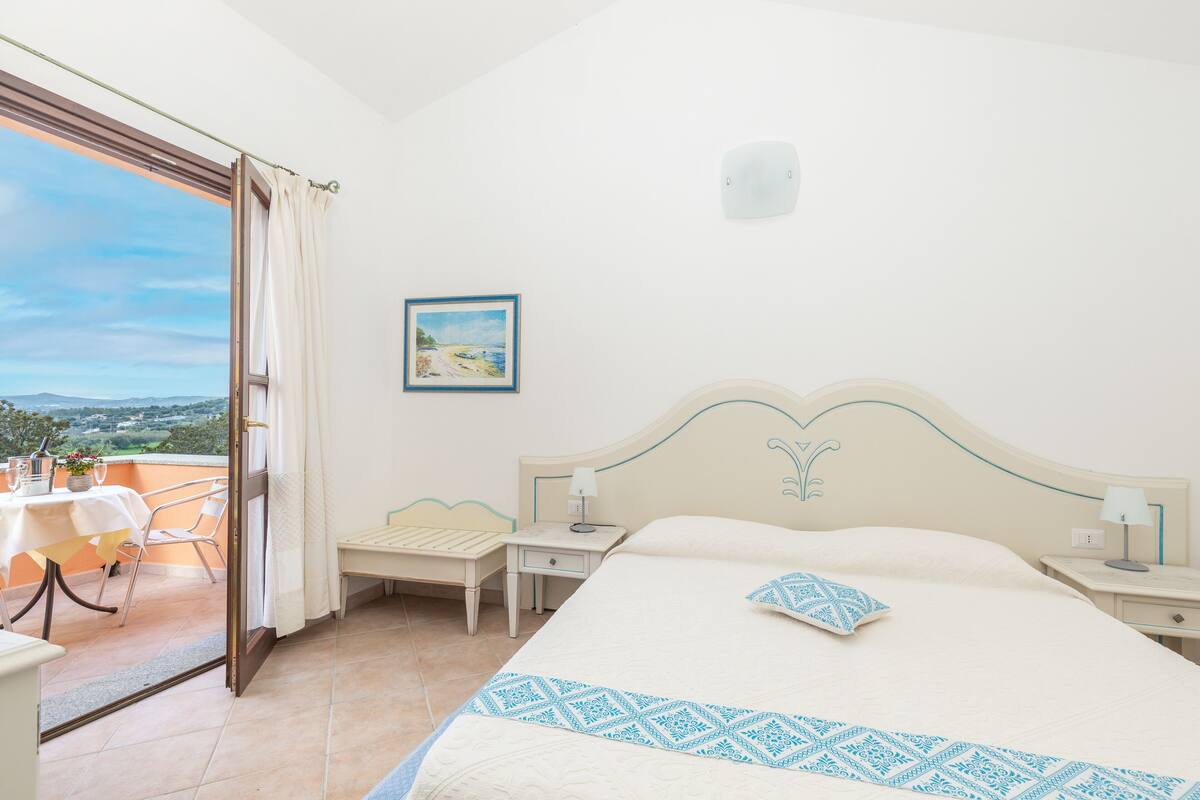 A bedroom with a bed, bedside tables, a desk, and a chair. There is a window with a view of the outdoors.