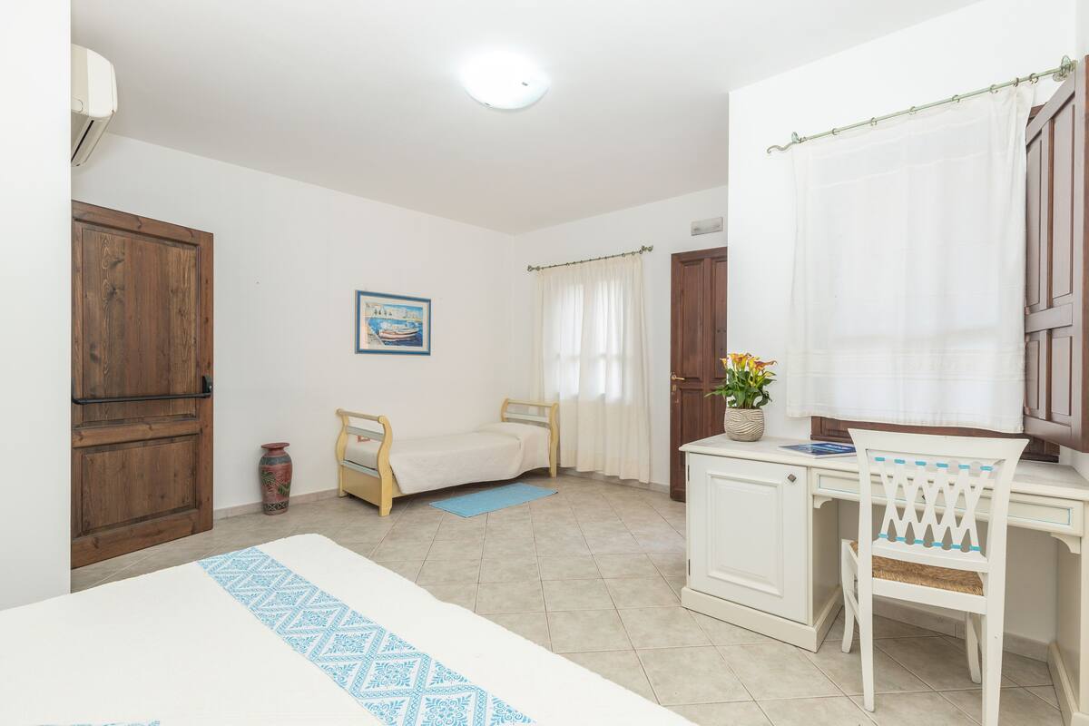 A bedroom with a bed, a desk, a chair, a wooden wardrobe, a small rug, a picture on the wall, and a window with curtains.