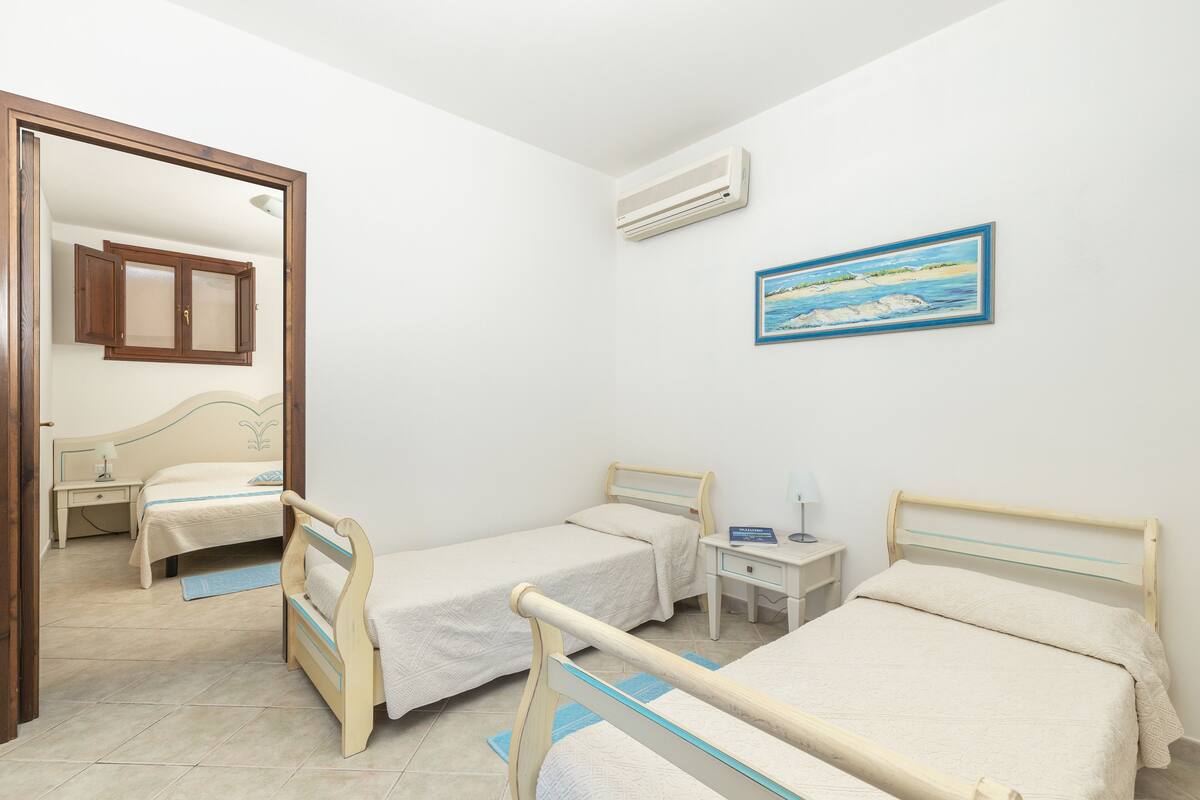 A bedroom with two single beds, a bedside table with a lamp, a small desk, and a wall-mounted air conditioner.