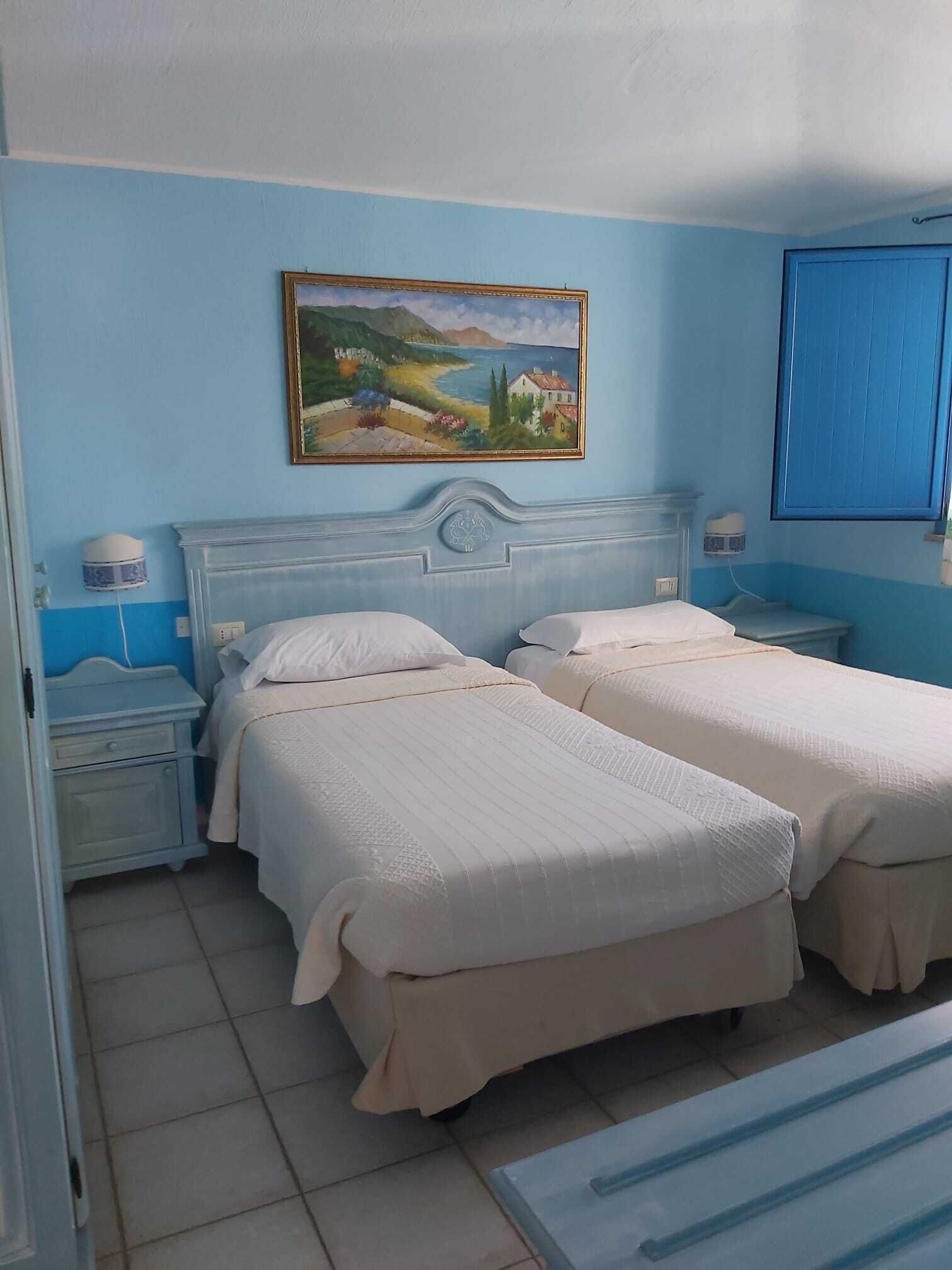 Un dormitorio con dos camas, una mesita de noche, una lámpara, un cuadro en la pared y una ventana con contraventanas azules.