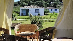 A view of two white buildings with tiled roofs, surrounded by greenery and hills. There are wicker chairs and a table in the foreground. - Resort Baia Cea (Bari Sardo)