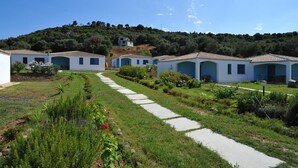 A row of white buildings with blue accents, a paved pathway, and a hillside in the background. - Resort Baia Cea (Bari Sardo)