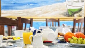 A breakfast table with a glass of orange juice, a white teapot, a plate with fruit, and a painting of a beach scene in the background. - Resort Baia Cea (Bari Sardo)