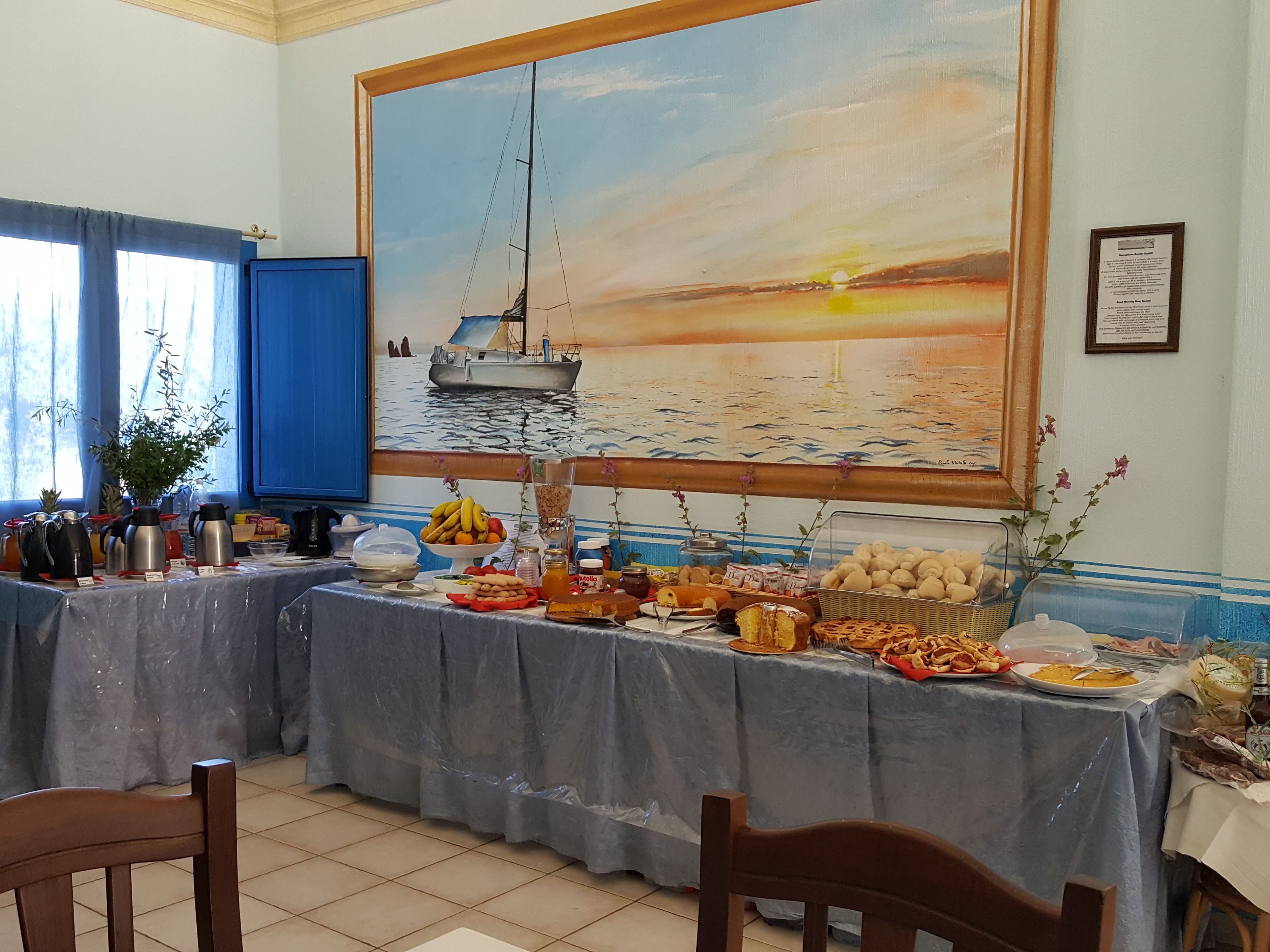 Un espace repas agrémenté d’un grand tableau représentant un coucher de soleil sur l’eau, d’une table de buffet où sont disposés divers plats, et d’un buffet bleu.