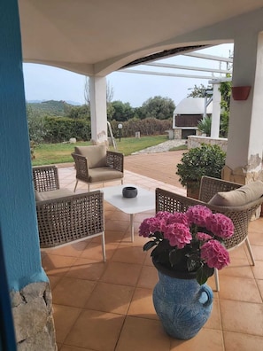Une terrasse extérieure avec des meubles en osier, une table blanche et un vase bleu contenant des fleurs roses.