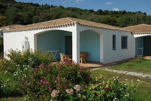 Un bâtiment blanc avec un toit recouvert de tuiles, deux fenêtres visibles et un petit espace extérieur aménagé avec des chaises.