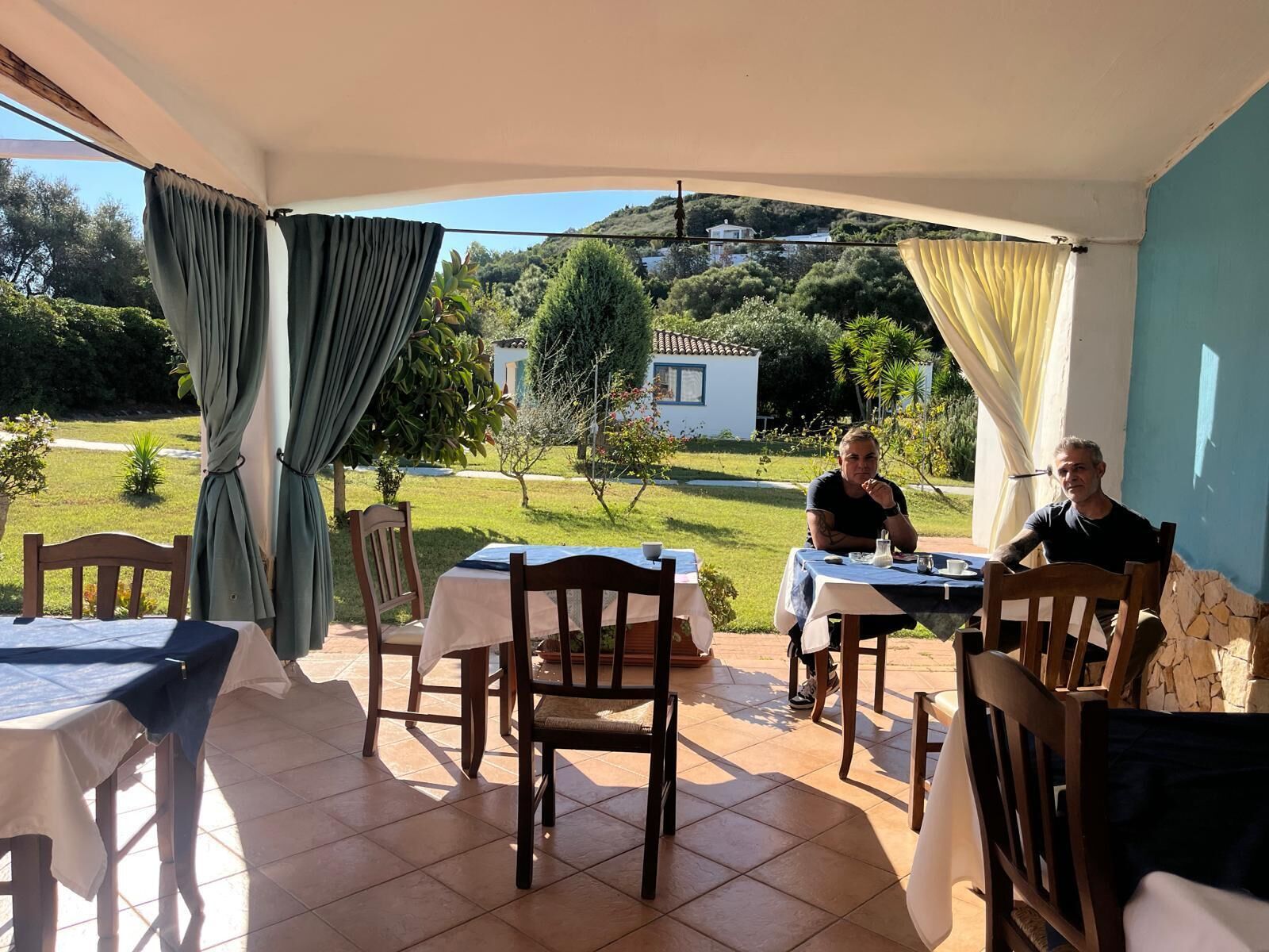 Un área de comedor al aire libre con techado, mesas y sillas, vista a una colina y un edificio en el fondo.