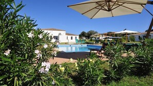 A pool area with lounge chairs and umbrellas, surrounded by greenery and a building in the background. - Resort Baia Cea (Bari Sardo)