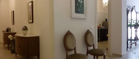 A hallway with a wooden cabinet, two upholstered chairs, a framed picture on the wall, and a chandelier in the background.