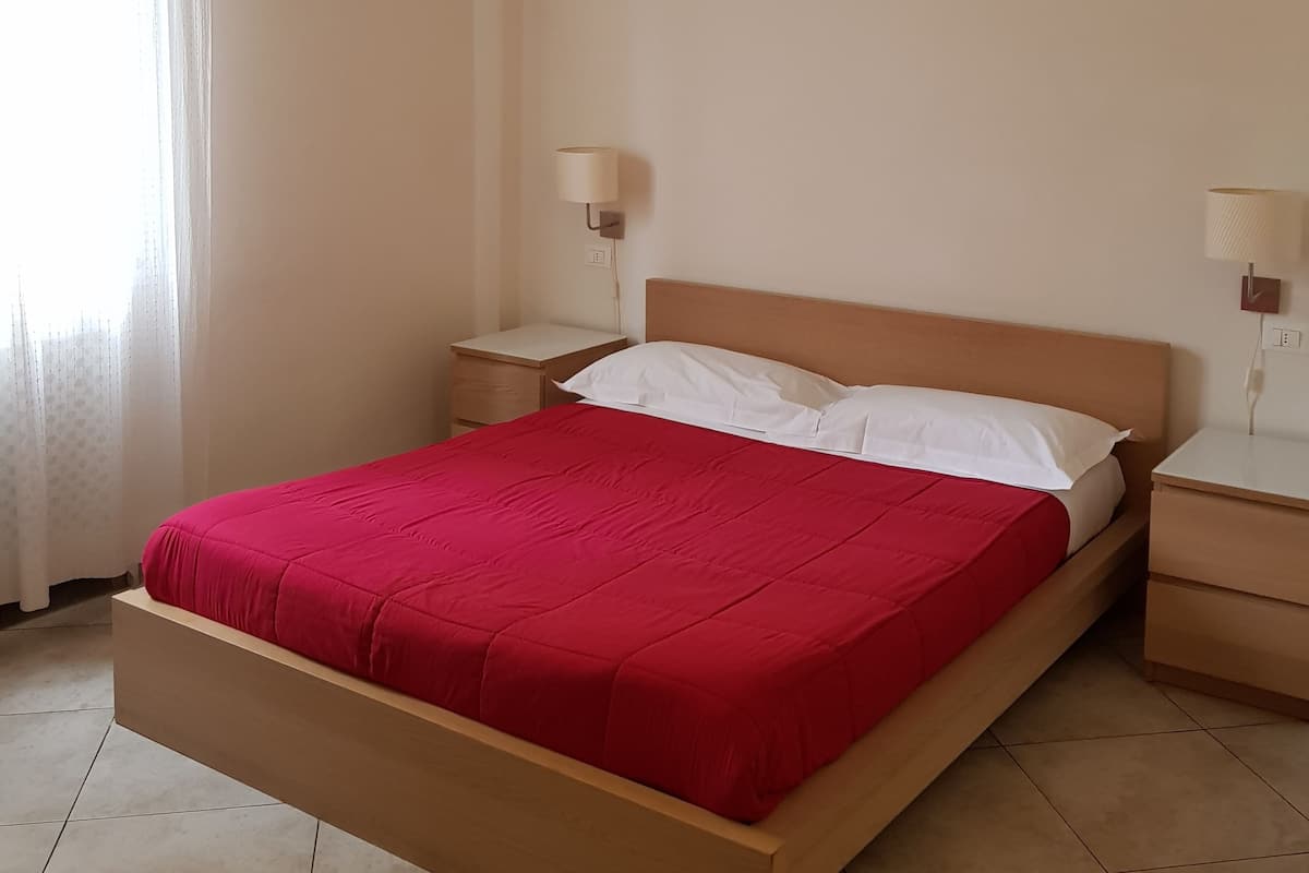 A neatly made bed with a red bedspread, two bedside tables with lamps, a window with sheer curtains, and a tiled floor.