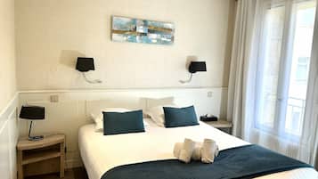 A hotel room with a bed, two bedside lamps, a wall art piece, a window with curtains, and a small bedside table with a lamp.