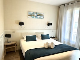 Habitación de hotel con cama, dos lámparas de noche, una obra de arte en la pared, una ventana con cortinas y una mesita de noche con lámpara.
