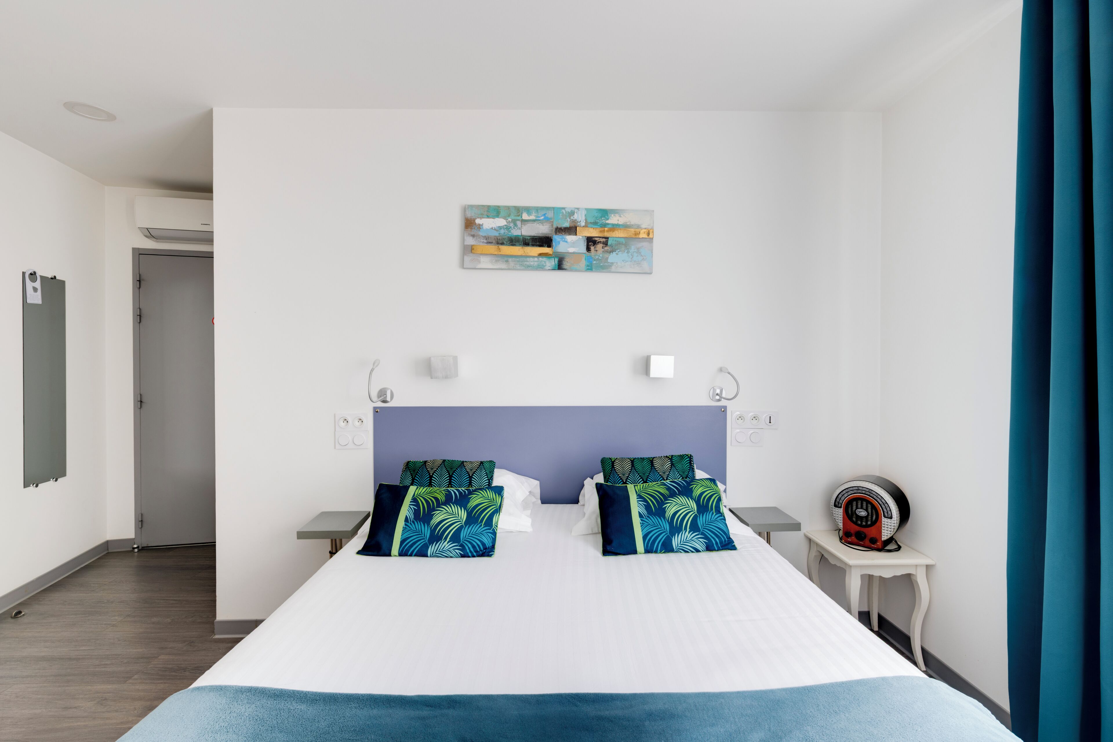 A hotel room with a bed, a bedside table, a wall-mounted radio, and a door leading to another room.