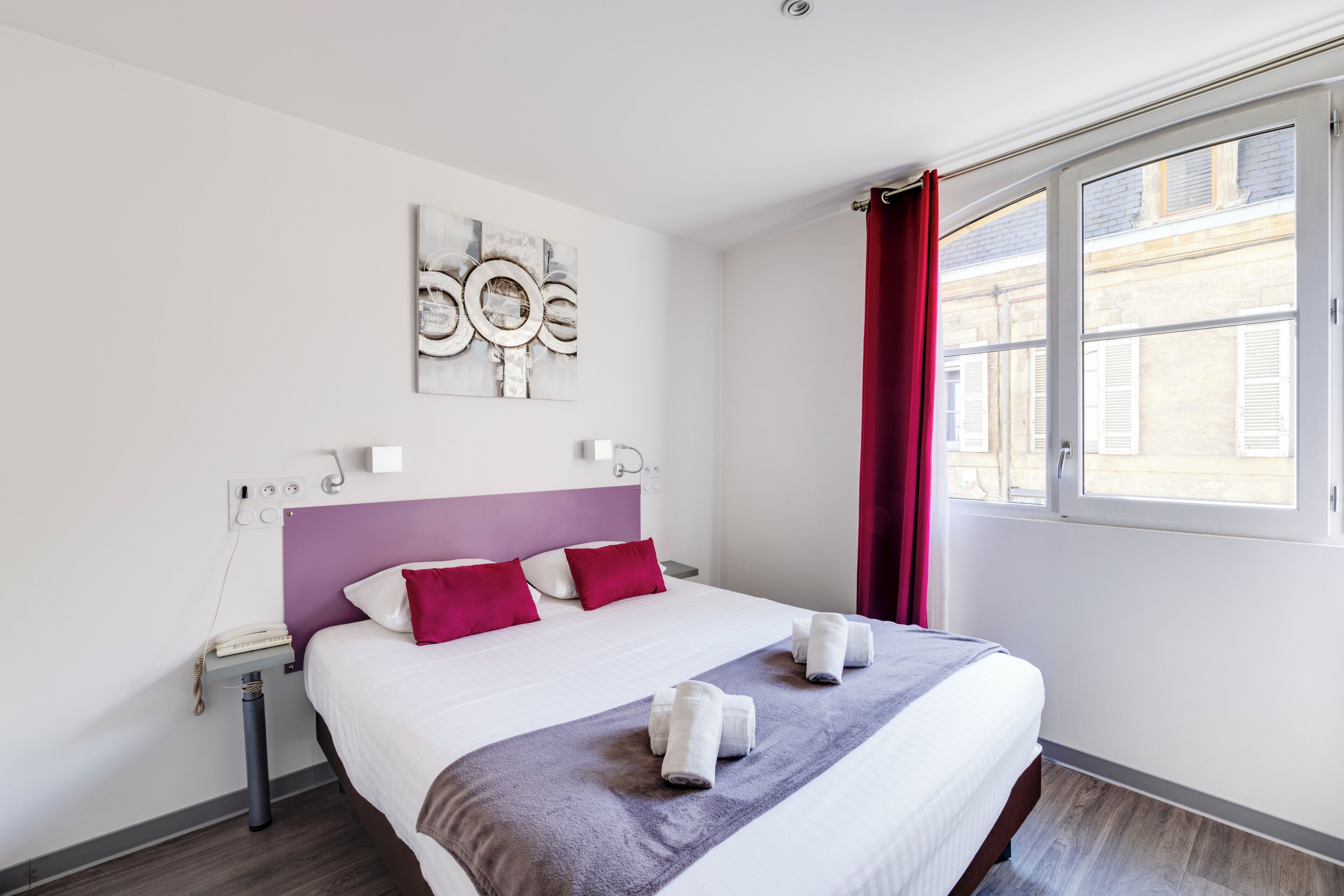 A hotel room with a bed, a bedside table, a telephone, a wall art piece, and a window with curtains.