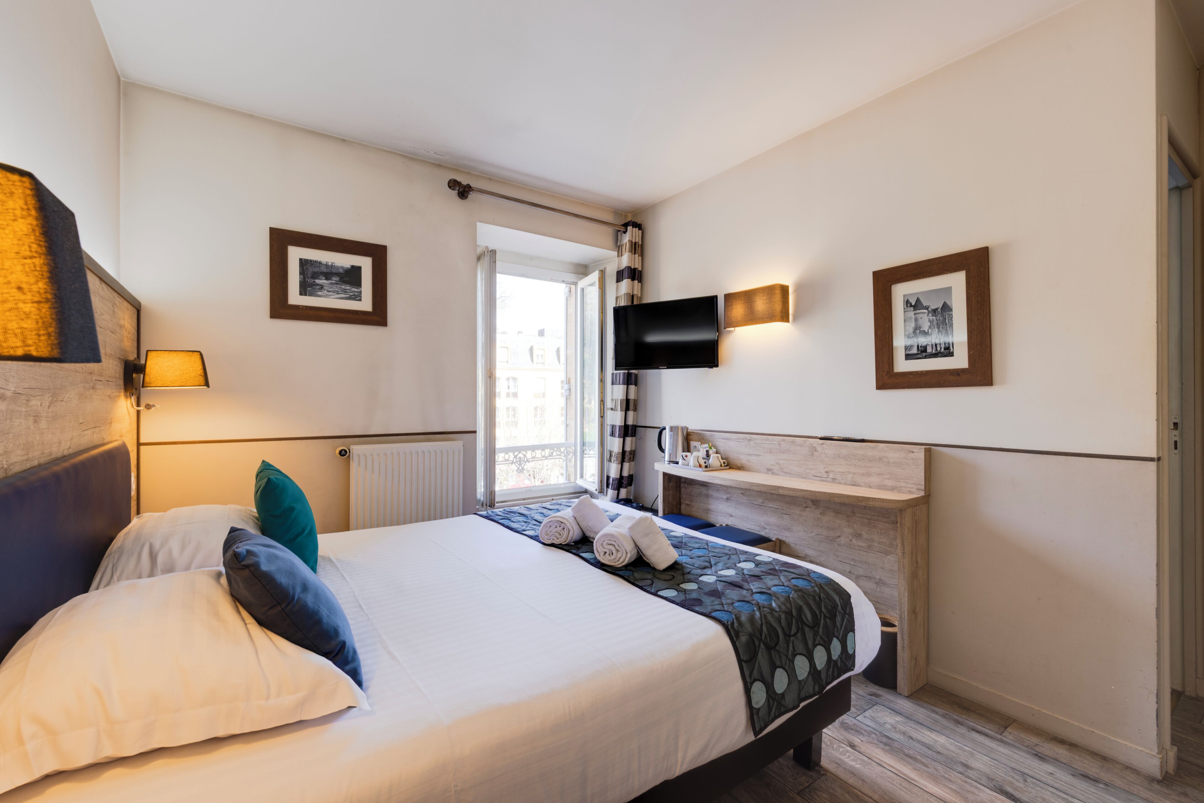 Une chambre d’hôtel avec un lit, des oreillers, une télévision, une fenêtre avec des rideaux et une étagère fixée au mur, sur laquelle sont posés divers objets.