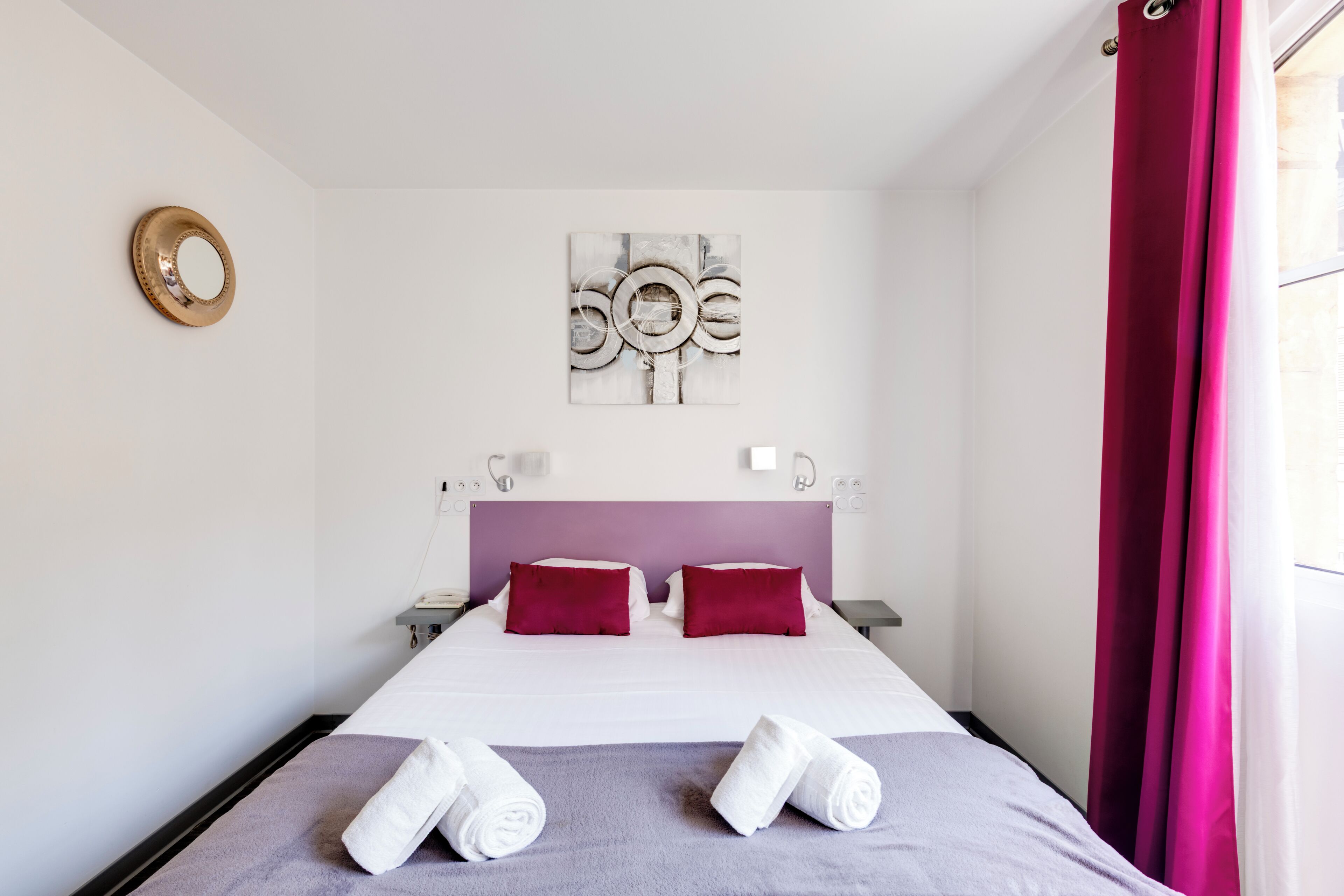 Una camera d'albergo con un letto, due cuscini, una testiera, un'opera d'arte appesa al muro, un comodino, una lampada, uno specchio e una finestra con tende.