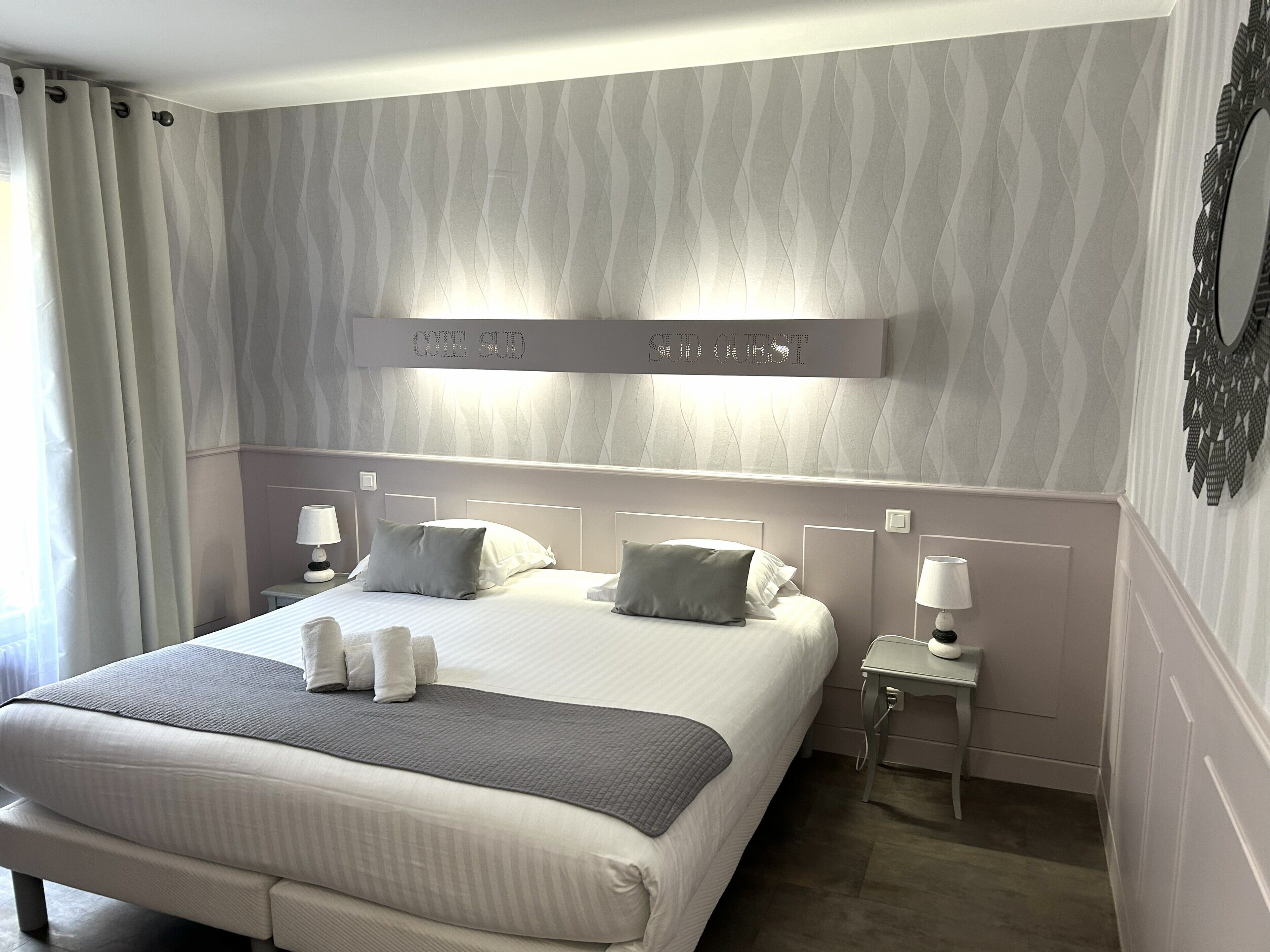 A hotel room with a large bed, two bedside tables, a headboard with a lighted sign, and a patterned wall.