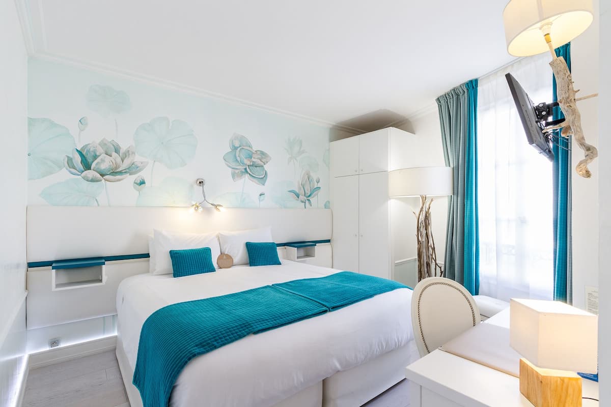 A modern bedroom with a large bed, a desk, and a chair. There is a wall with a floral mural, a ceiling light, and a window with curtains.