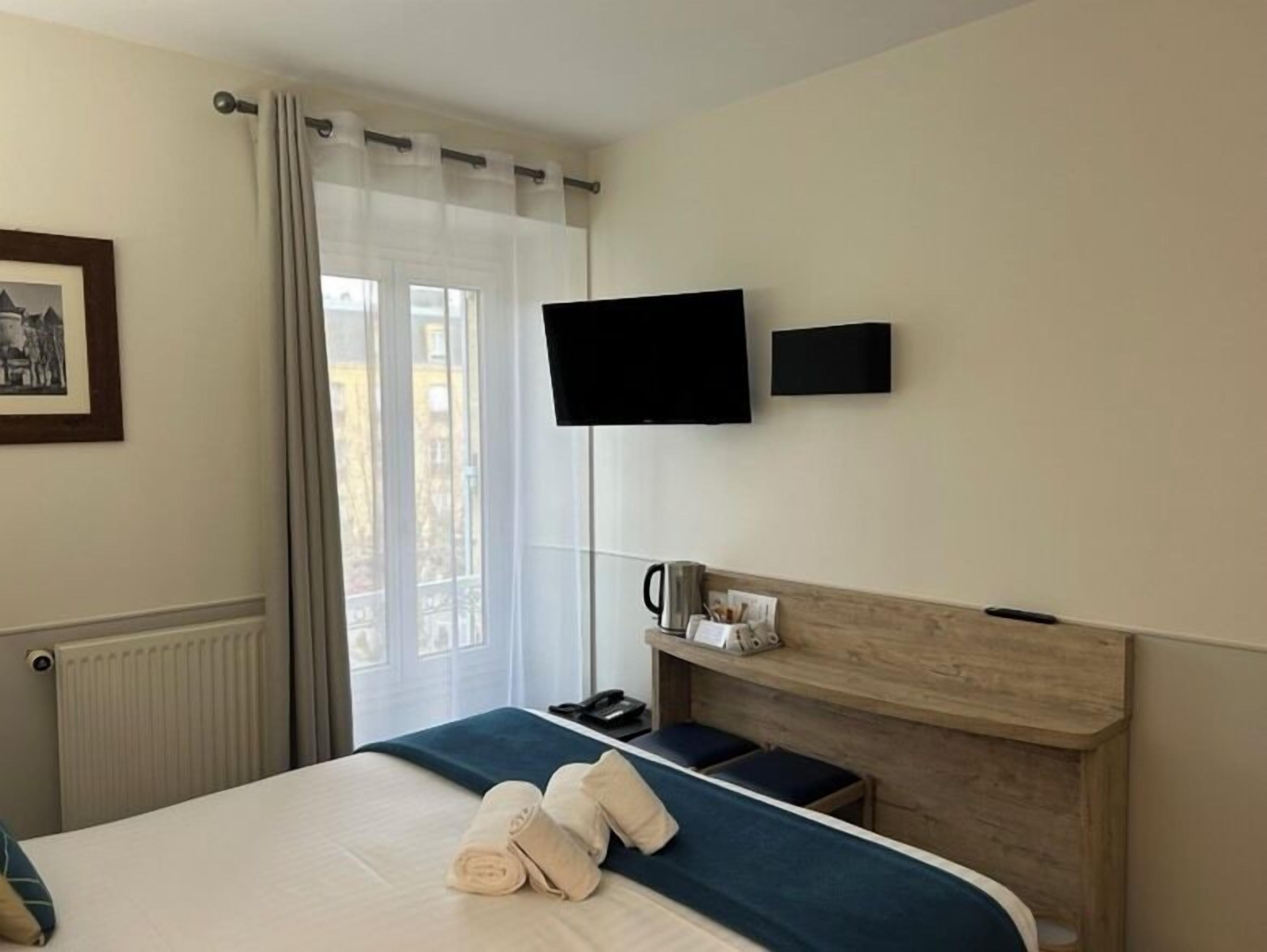 A hotel room with a bed, a flat-screen TV mounted on the wall, a bedside table with a lamp, and a window with curtains.