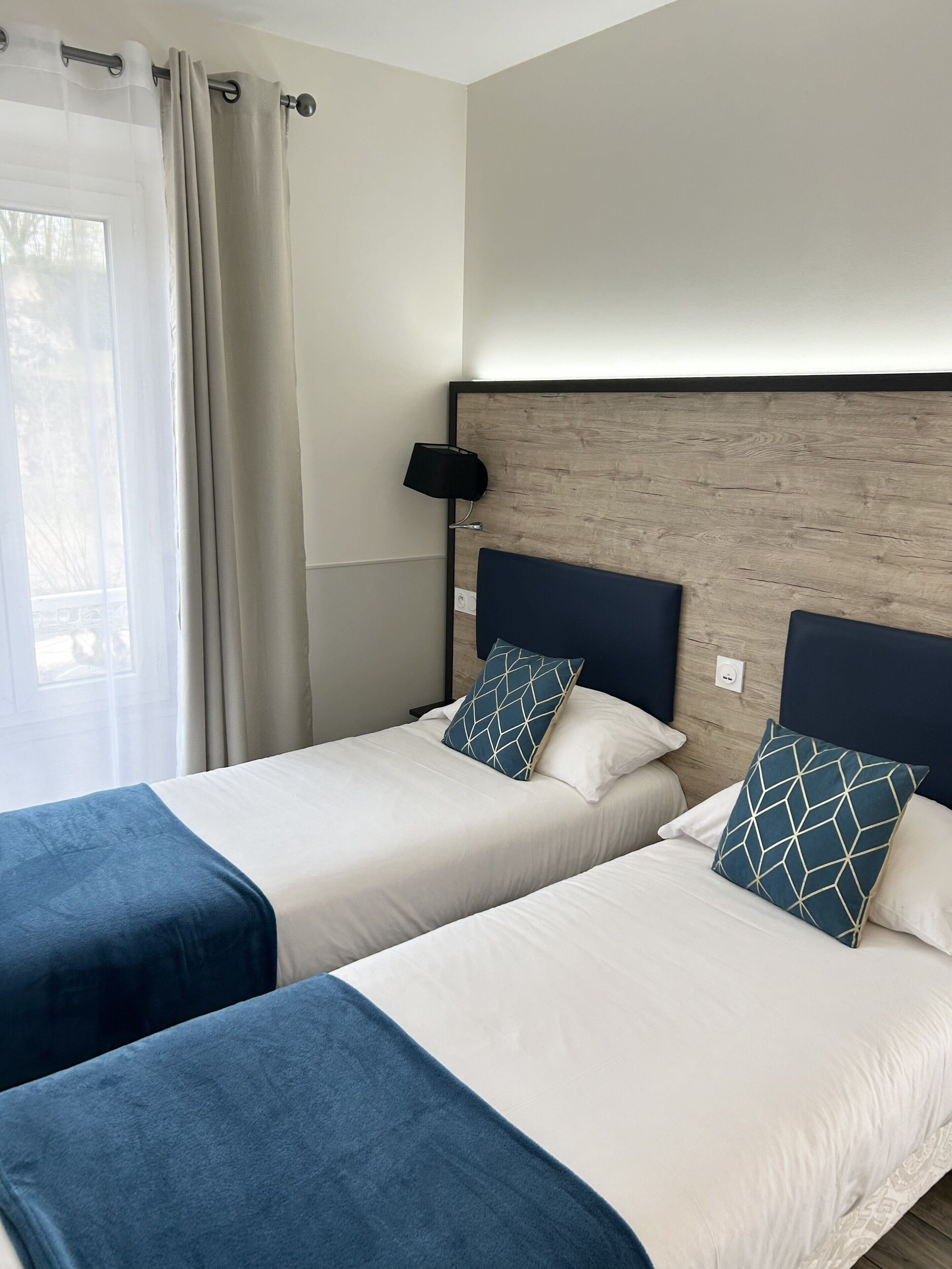 A hotel room with two beds, a wooden headboard, a bedside lamp, and a window with curtains.