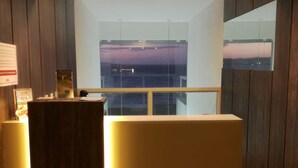 A reception desk with a wooden panel and a glass partition. There are three lights above the desk, and a view of a sunset through the glass. - Aparthotel Margherita (Natal)