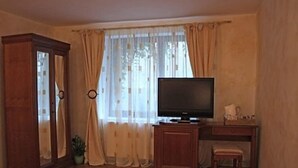 Una habitación con un armario de madera, un televisor sobre un mueble bajo, un escritorio con una silla y una ventana con cortinas traslúcidas.