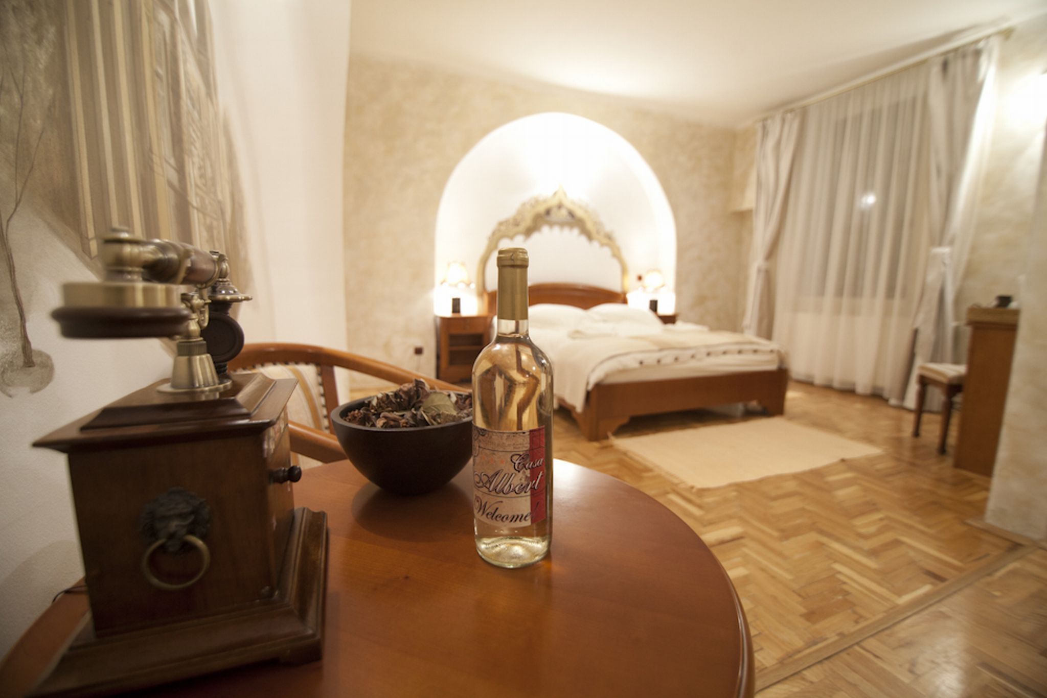 A hotel room with a wooden table, a bottle of wine, a vintage stove, a bed with a canopy, and a nightstand with a lamp.