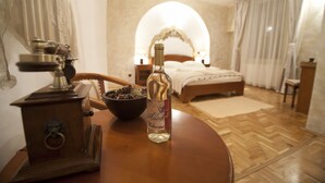 Una habitación de hotel con una mesa de madera, una botella de vino, una estufa antigua y una cama con dosel, además de una mesita de noche con una lámpara.
