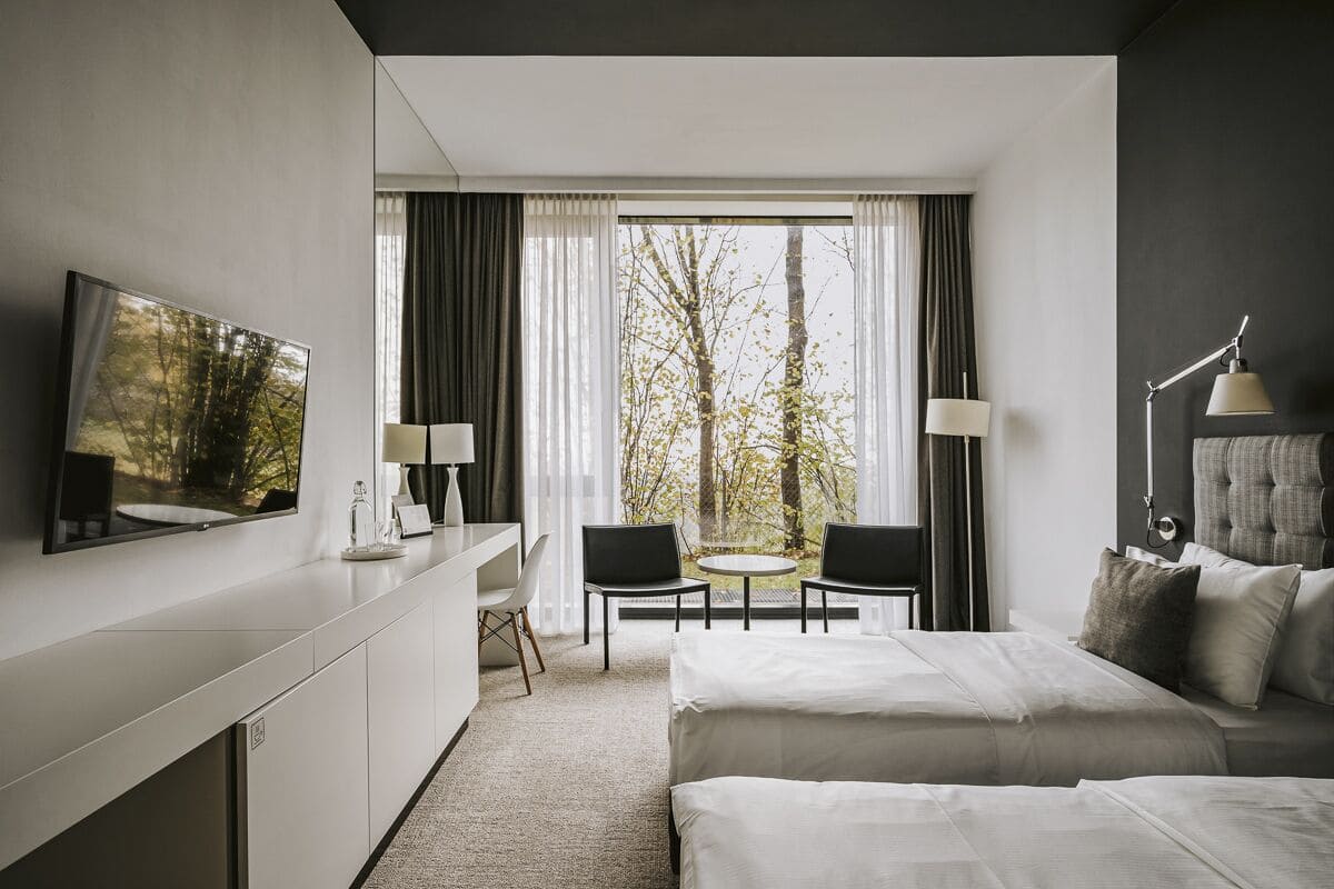 Ein modernes Hotelzimmer mit Bett, Schreibtisch, Stuhl, Fernseher und Blick auf Bäume durch große Fenster.