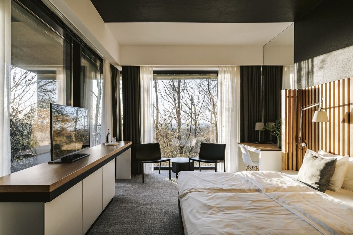 Ein modernes Hotelzimmer mit einem großen Bett, einem Schreibtisch mit Fernseher und Blick auf Bäume durch das Fenster.