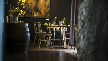 Un comedor con mesa de madera dispuesta para dos comensales, sillas, un barril grande y un cuadro en la pared.