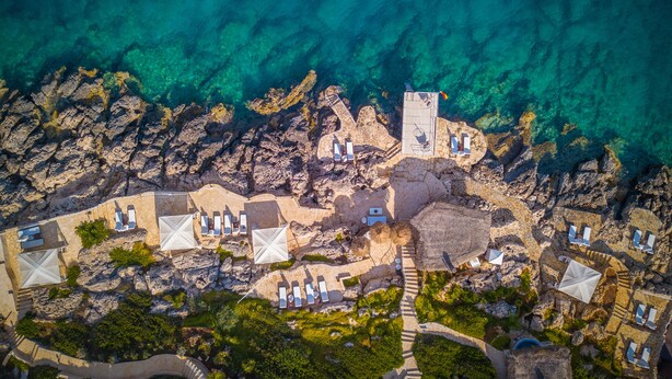 An aerial view of a coastal resort with clear blue waters, rocky terrain, and multiple lounge areas with umbrellas. - Lukka Exclusive Hotel - Adult Only +12 (Kas)
