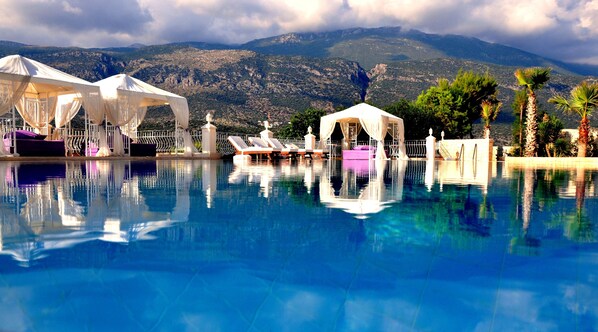 A pool with white canopies and lounge chairs, surrounded by palm trees and mountains in the background. - Lukka Exclusive Hotel - Adult Only +12 (Kas)