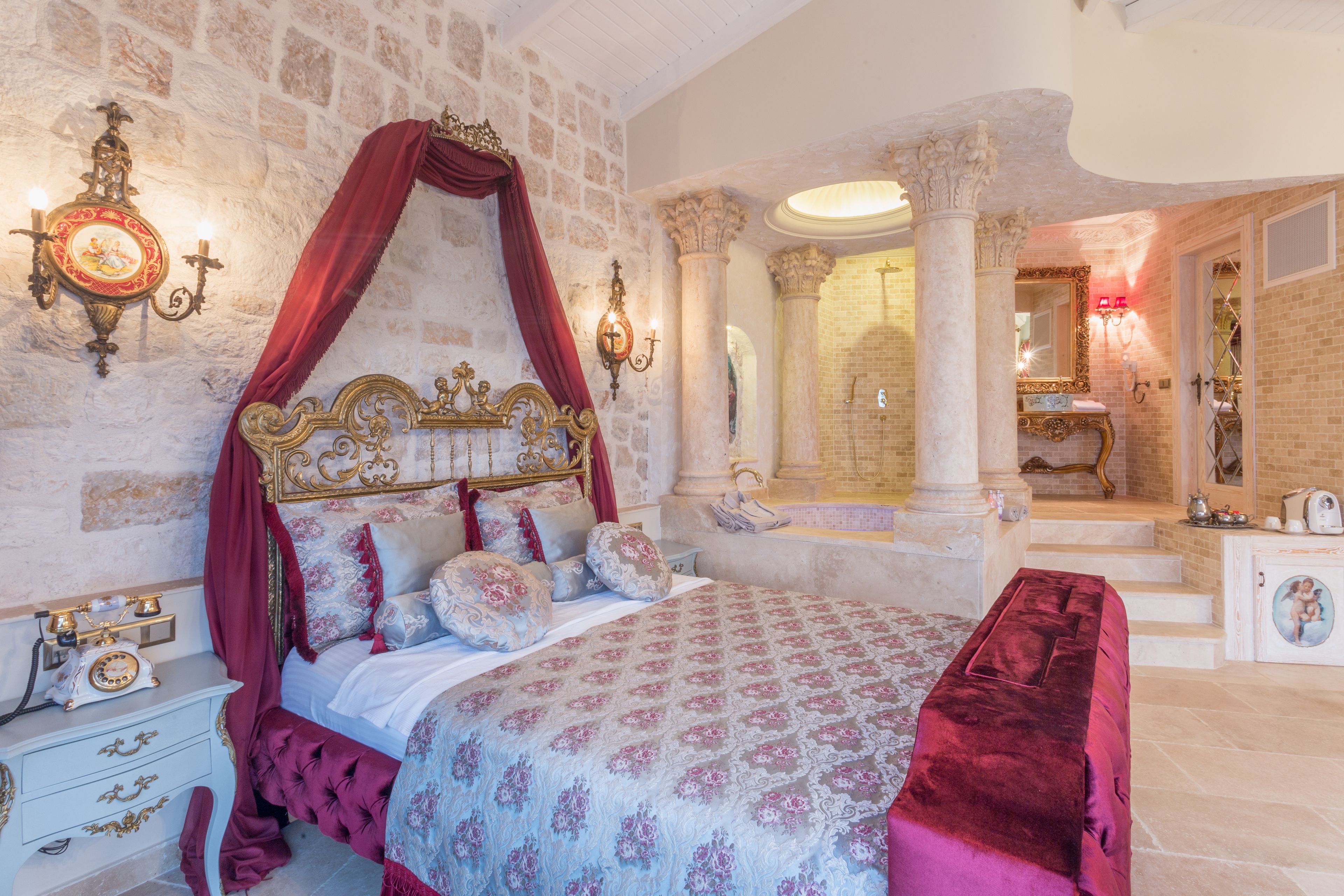 A luxurious bedroom with a four-poster bed, a marble column, a large mirror, and a vanity with a bathtub.