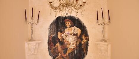 A room with a large oval painting on the wall, a chandelier, and candelabras.